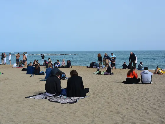 La playa de la Barceloneta en un día soleado, con gente sentada en la arena en grupos pequeños y el mar al fondo.