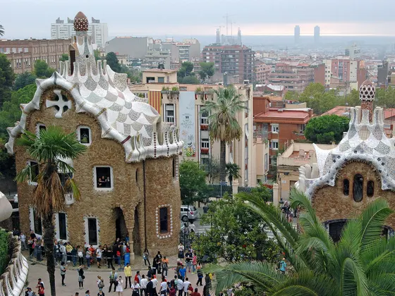 Los dos pabellones de la entrada del Park Güell, con sus tejados de trencadís blanco y azul, palmeras delante y la ciudad extendida al fondo hasta el horizonte.