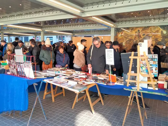 El mercado de libros de Sant Antoni bajo el tejadillo del mercado, con mesas llenas de libros y cómics, un caballete con carteles y gente mirando los puestos.