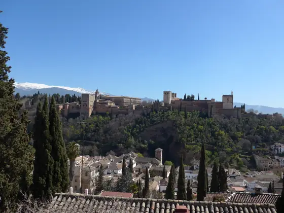 La Alhambra sobre la colina, con Sierra Nevada nevada al fondo, vista por encima de los tejados y los cipreses del Albaicín.