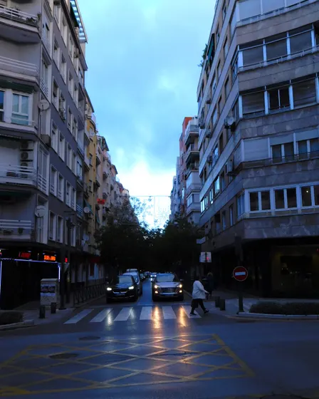 Calle Pedro Antonio de Alarcón al anochecer, con bloques de pisos a los dos lados, coches con los faros encendidos y un paso de cebra en primer plano.