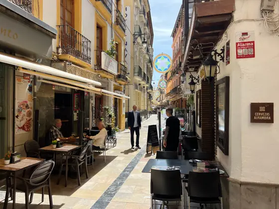 Terrazas y mesas altas a los dos lados de una calle estrecha de Granada, con gente tomando algo a media tarde.