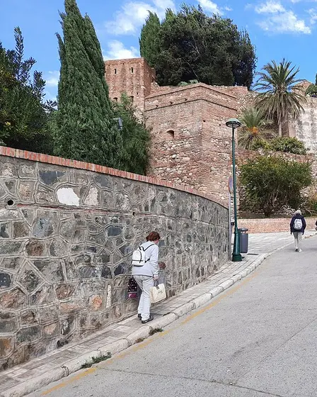 La muralla de ladrillo y piedra de la Alcazaba entre cipreses y palmeras, con una calle en cuesta junto al muro por la que suben dos personas.