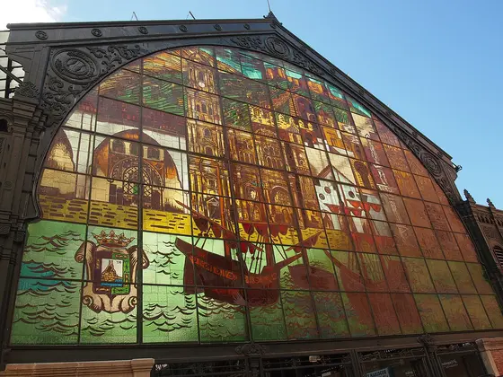La gran vidriera de colores de la fachada del mercado de Atarazanas, con el escudo de Málaga, un barco y edificios de la ciudad, bajo un cielo azul.