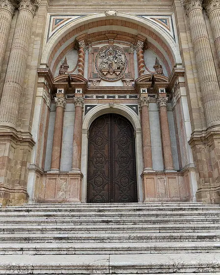 La portada principal de la Catedral de Málaga, de mármol rojizo, con columnas a los lados, un escudo tallado sobre la puerta de madera y una escalinata de piedra delante.