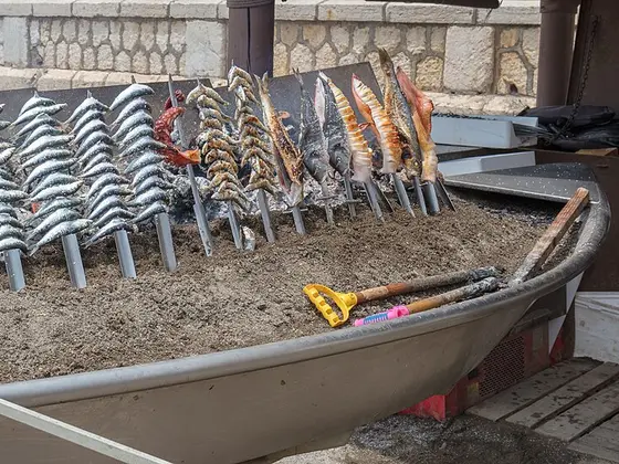 Una barca varada llena de arena con espetos de sardinas y otros pescados clavados en cañas, junto a un muro de piedra.