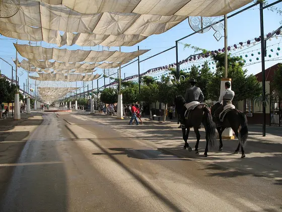 Una calle ancha del recinto de la Feria de día, cubierta con toldos blancos y guirnaldas de farolillos, con dos jinetes a caballo a la derecha.