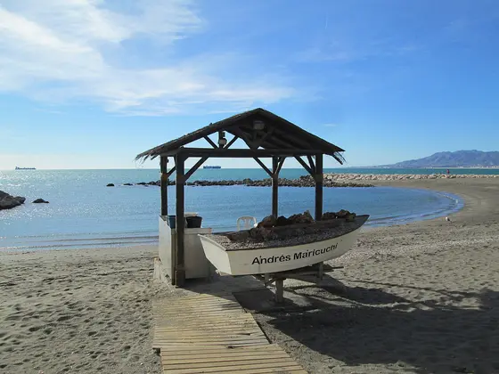 La orilla de Pedregalejo con una barca de pesca varada en la arena bajo un cobertizo de madera, y el mar en calma bajo un cielo azul.