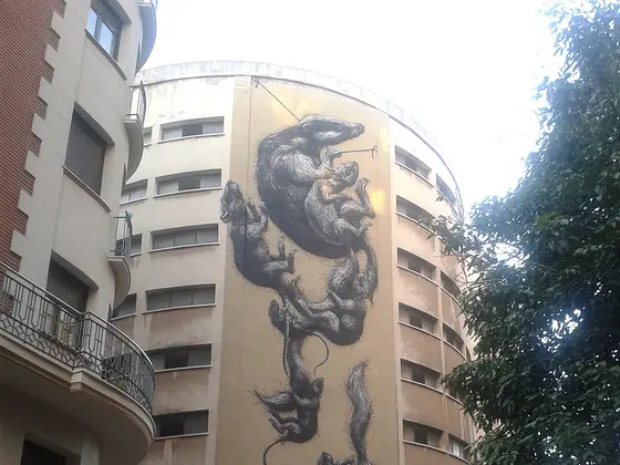 Un edificio de esquina redondeada con la fachada entera pintada con ratas gigantes en gris, colgadas cabeza abajo, junto a un balcón y las ramas de un árbol.