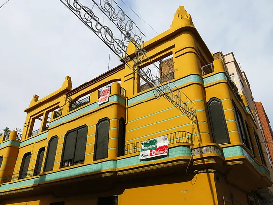 Una casa de esquina del Cabanyal de dos plantas, pintada de amarillo con molduras y franjas verde agua, vista desde abajo contra un cielo gris.