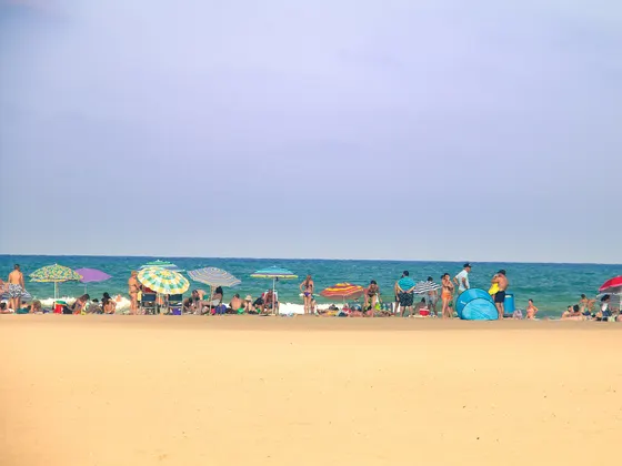La playa de la Malvarrosa en un día de verano, con arena ancha en primer plano, sombrillas de colores y bañistas en la orilla y el mar azul al fondo.