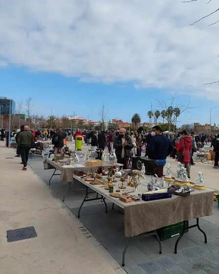 El Rastro en un paseo ancho con árboles jóvenes, mesas plegables llenas de objetos viejos a la derecha y mucha gente entre los puestos.