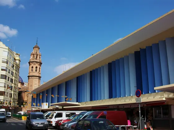 El mercado de Ruzafa, un edificio largo con la fachada de lamas verticales azules, y detrás el campanario de una iglesia bajo un cielo azul.