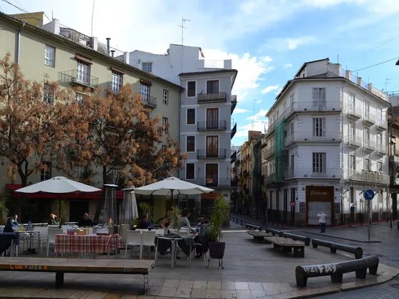 La plaza del Tossal con mesas y sombrillas de terraza, bancos de piedra, árboles de hojas secas y edificios de cinco plantas alrededor.