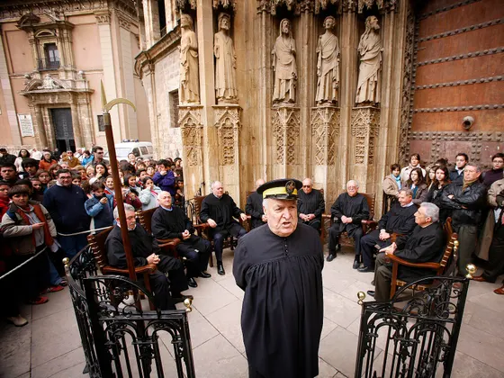 Una sesión del Tribunal de las Aguas ante la portada gótica de la Catedral: los síndicos vestidos de negro sentados en sillones, un alguacil en primer plano y el público detrás de una verja.